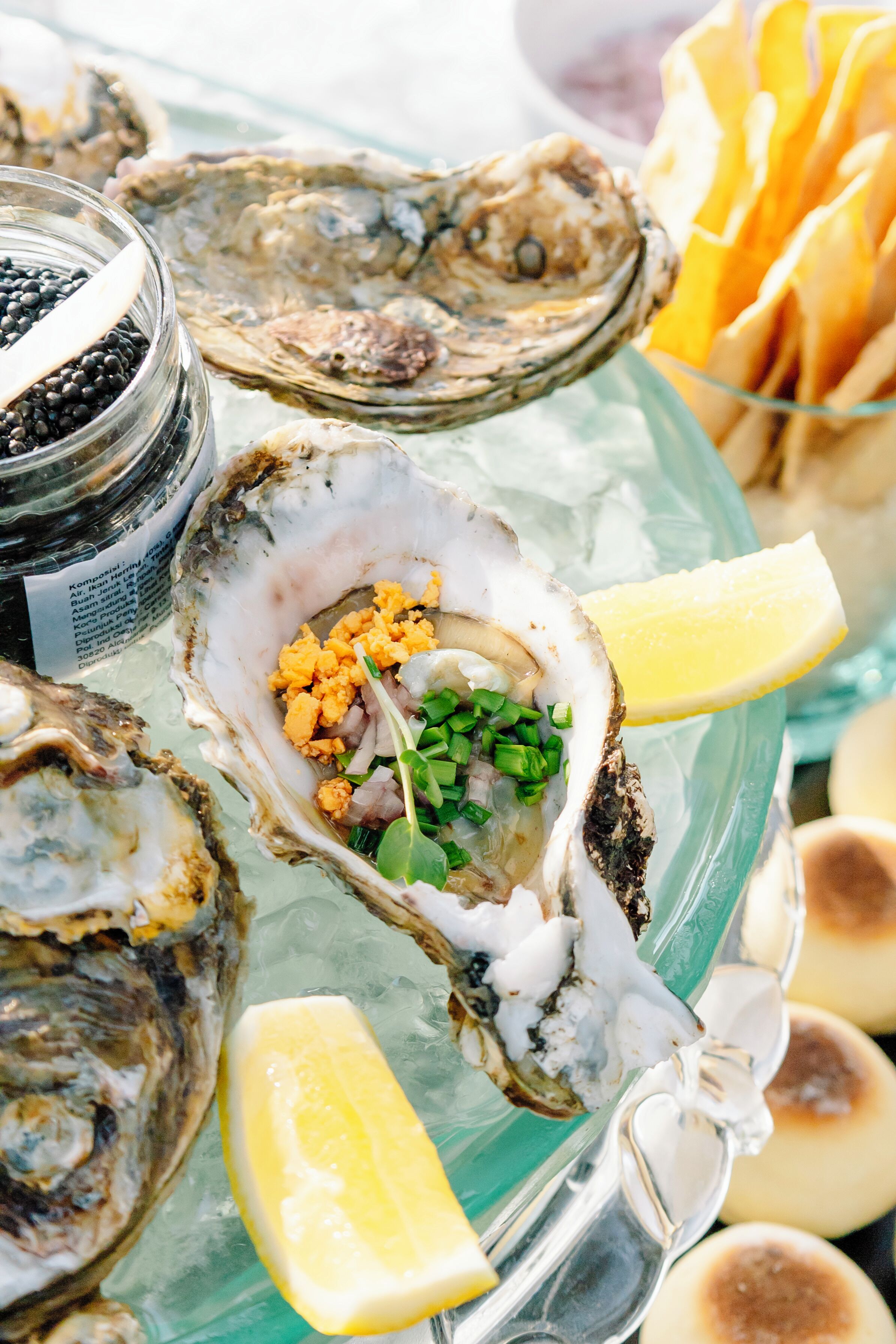 A tray of oysters on ice with lemon wedges and a jar of black olives.
