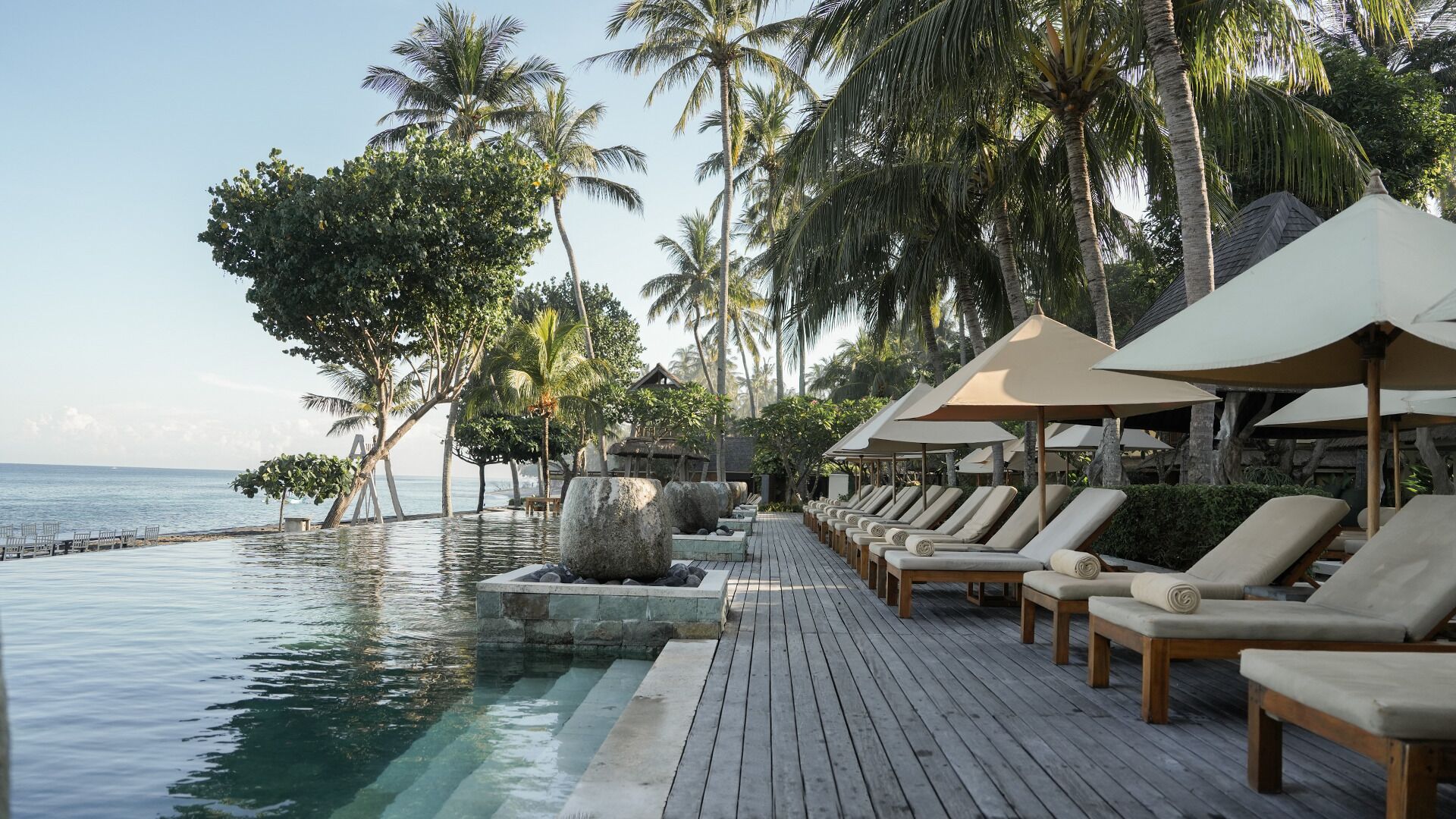 Une terrasse en bois, dotée de chaises longues et de parasols, surplombe une piscine, le tout étant entouré de palmiers et offrant une vue sur la mer.