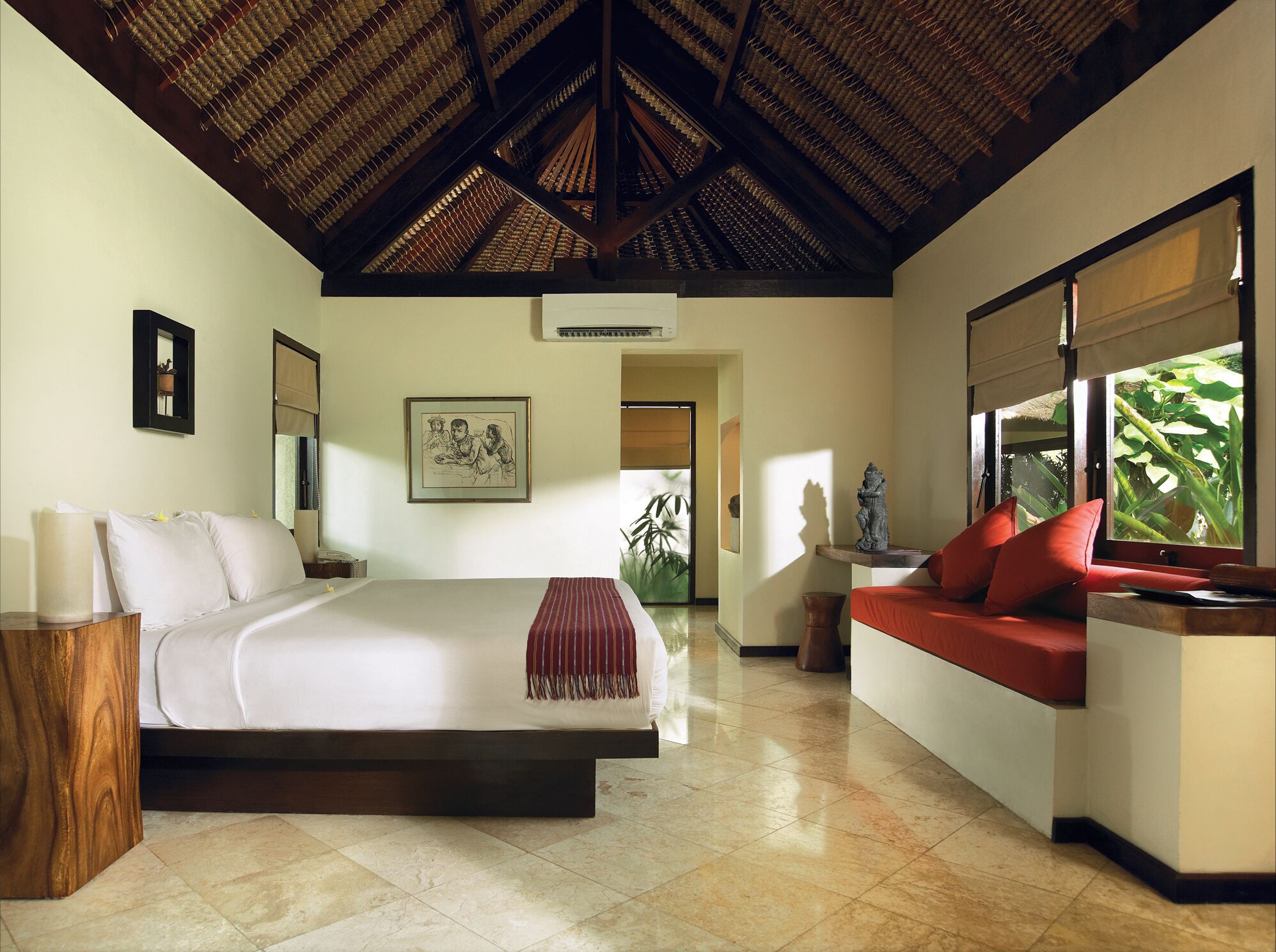 A spacious bedroom with a large bed, a wooden nightstand, a red sofa, and a window with a view of greenery.