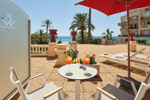A sunlit outdoor seating area with lounge chairs and a table, featuring a view of the sea and palm trees.
