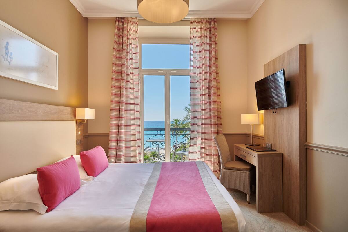 Habitación de hotel con cama, dos lámparas de noche, televisor montado en la pared y vista al océano a través de la ventana.