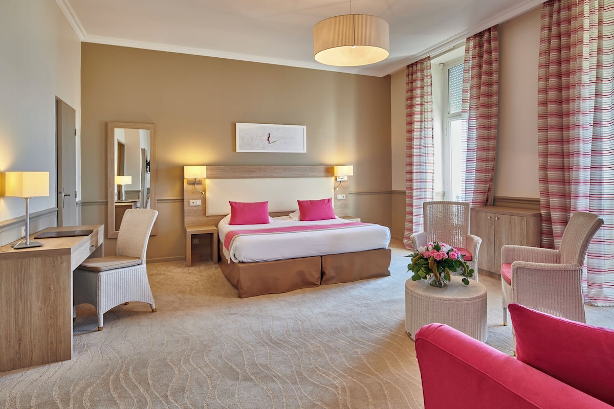 Habitación de hotel con cama, dos mesitas de noche con lámparas, un escritorio, una silla, una pequeña mesa redonda con flores y un ventanal con cortinas.