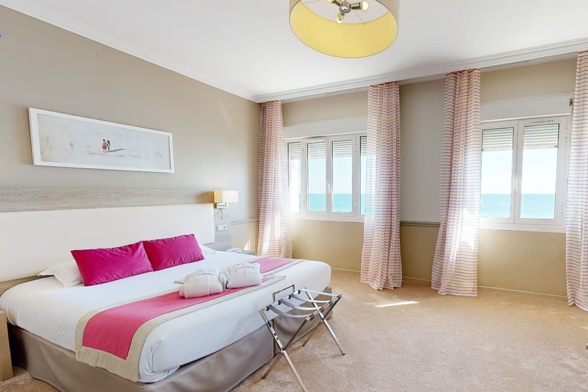 Una habitación de hotel con una cama, cabecero, cortinas en la ventana, ventilador de techo y un cuadro en la pared.