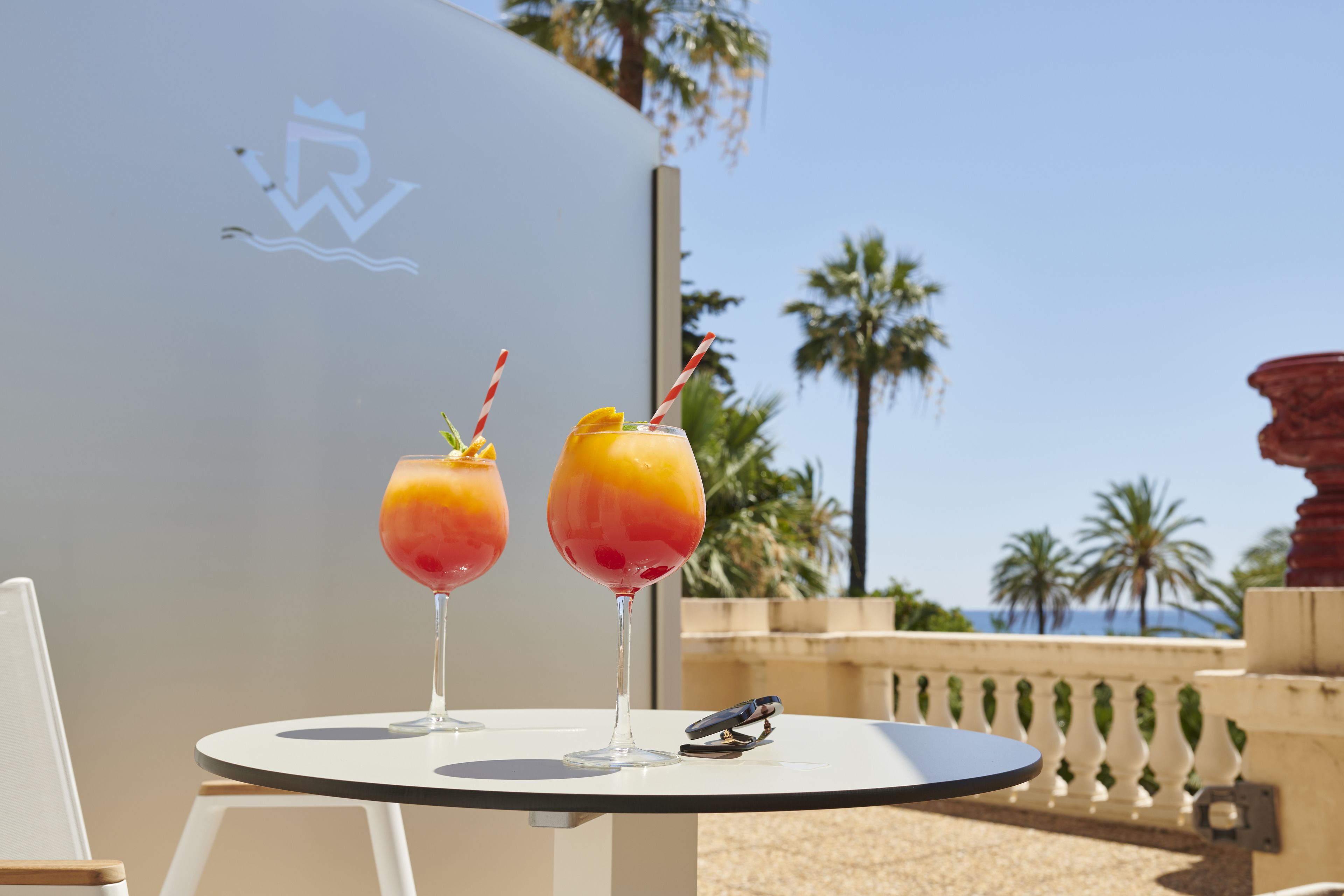 Two drinks on a table with a view of palm trees and the ocean.