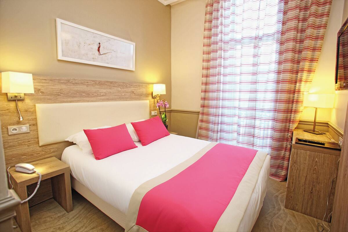 Una habitación de hotel con una cama grande, dos lámparas de noche, una mesita de noche con un teléfono, un reloj de pared y una ventana con cortinas a rayas.