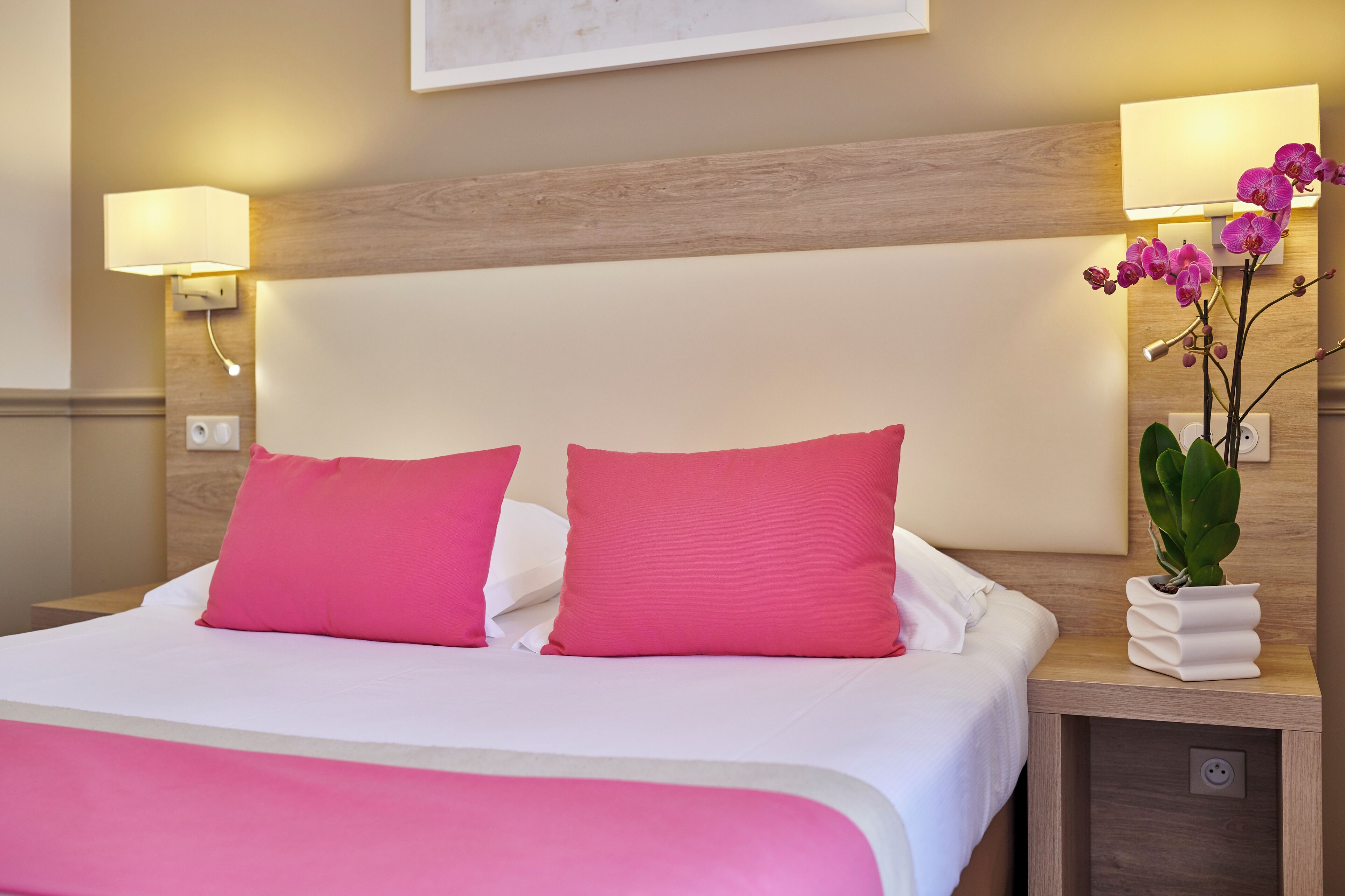 A hotel room with a large bed, two pink pillows, a bedside table with a lamp, and a potted plant.
