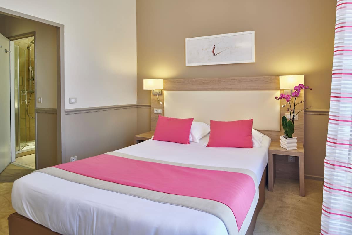 Habitación de hotel con una cama grande, dos mesitas de noche, una lámpara de pared, un cuadro enmarcado y una planta en maceta.
