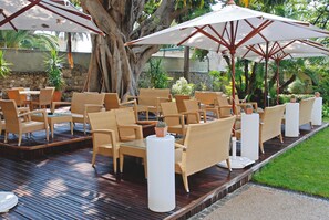 An outdoor seating area with wicker chairs and tables, shaded by umbrellas, and surrounded by greenery.