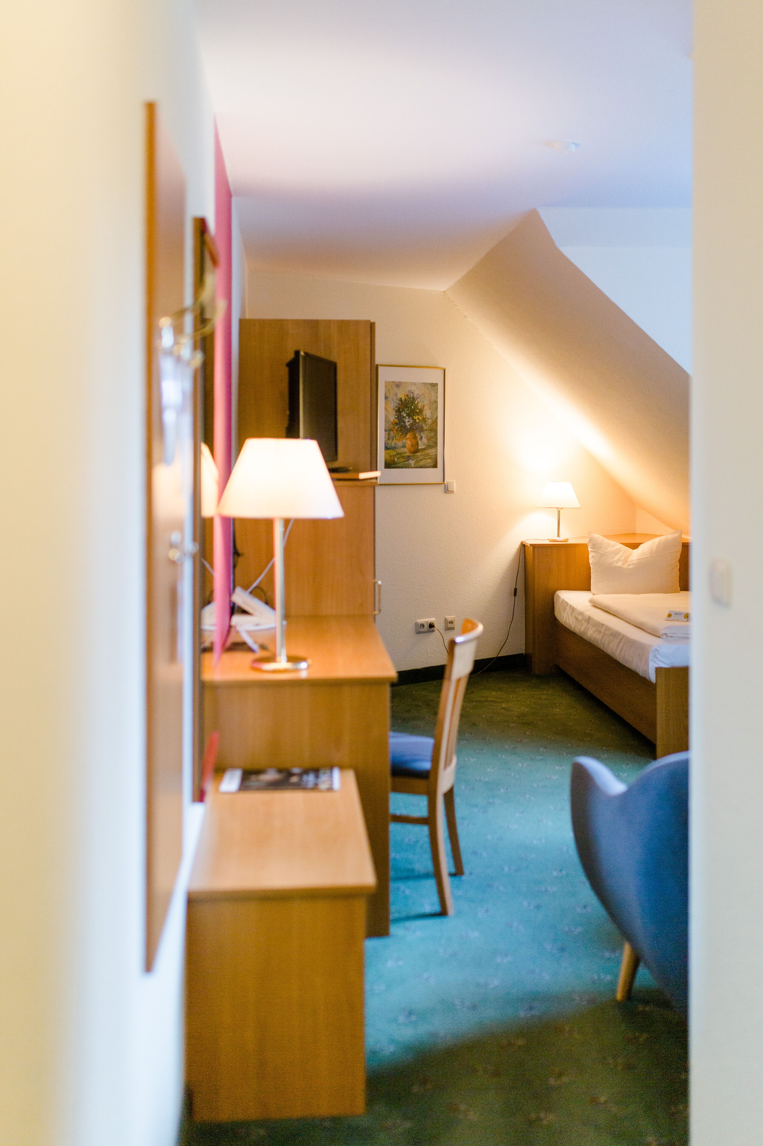A small, cozy room with a desk, chair, and a bed. There is a lamp on the desk and another on the bedside table.