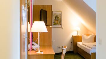 A small, cozy room with a desk, chair, and a bed. There is a lamp on the desk and another on the bedside table.