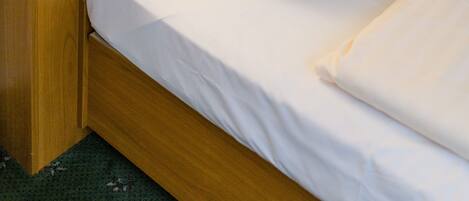 Un lit bien fait, avec du linge de lit blanc et un seul oreiller blanc, une table de chevet en bois avec une lampe, et une tête de lit en bois.