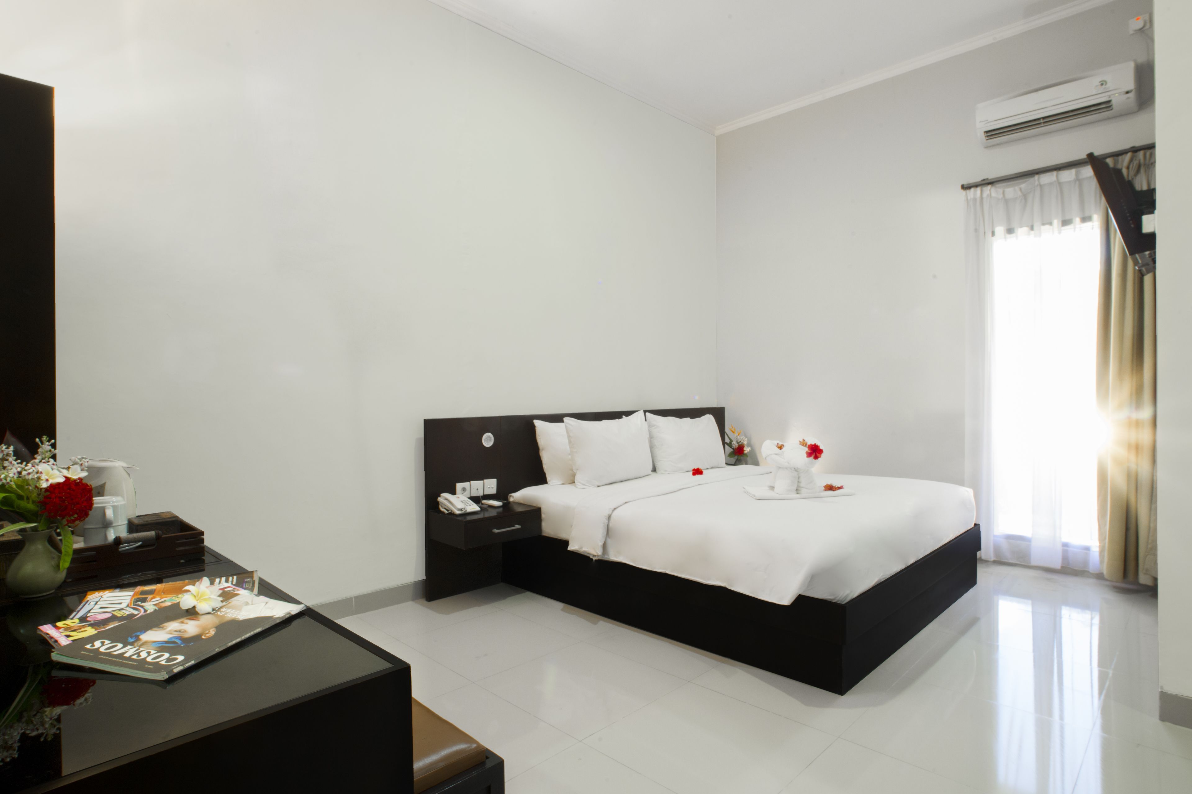 Een moderne hotelkamer met een groot bed, een nachtkastje met een tijdschrift, een vaas met bloemen en een raam met gordijnen.