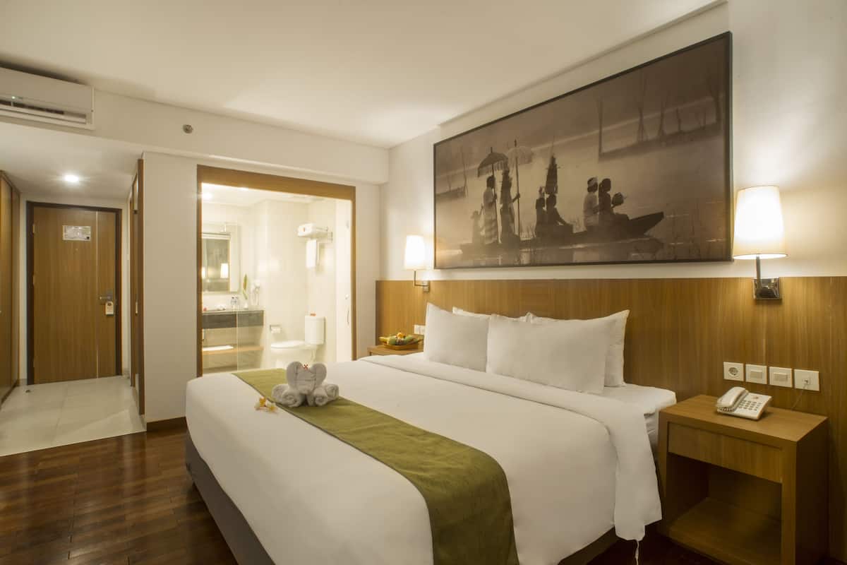 Hotellihuone, jossa on suuri sänky, puinen pääty, sängyn yläpuolella oleva taulu, yöpöytä, jossa on puhelin, ja ovi, joka johtaa kylpyhuoneeseen.