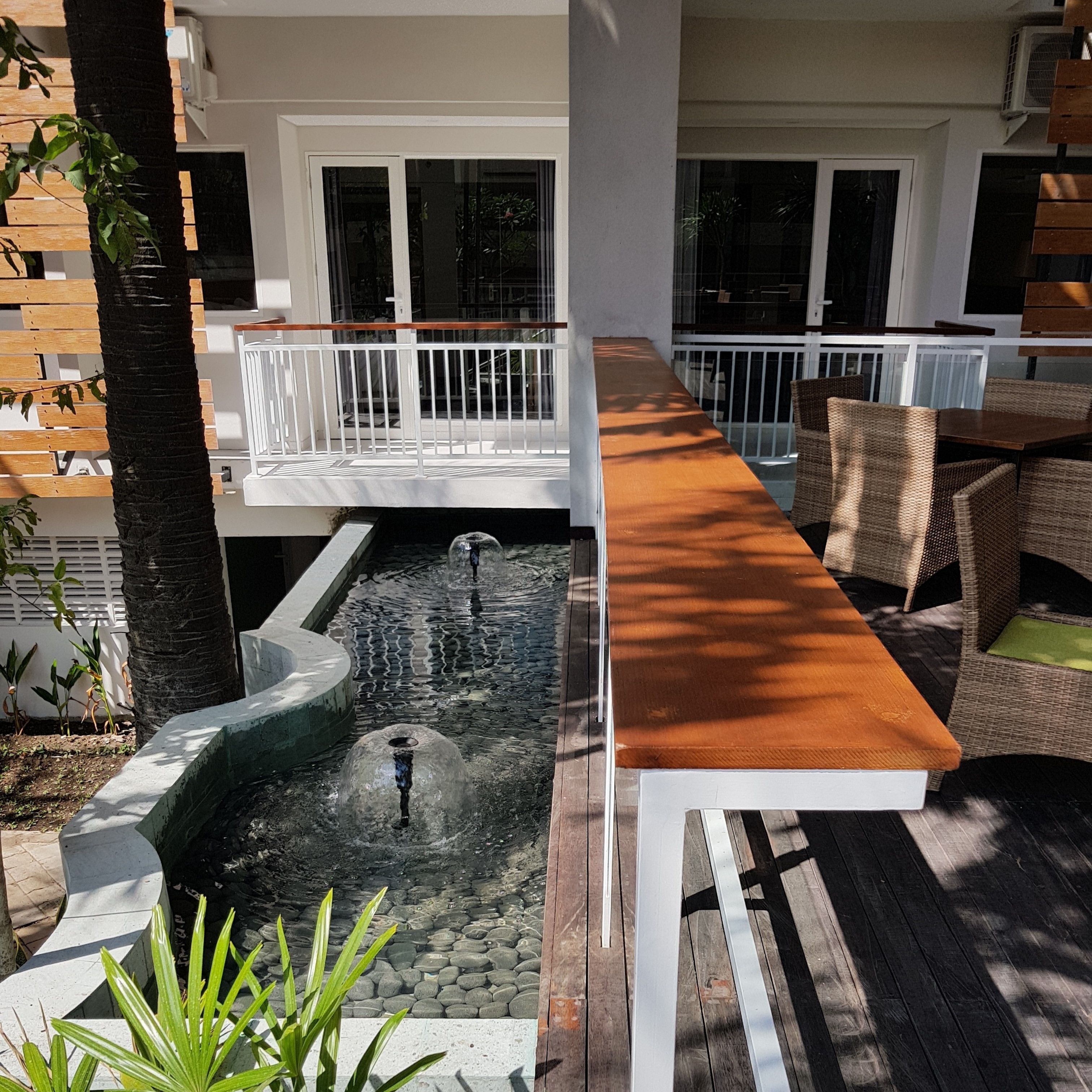 A modern outdoor deck with a long wooden table, wicker chairs, and a small water feature.