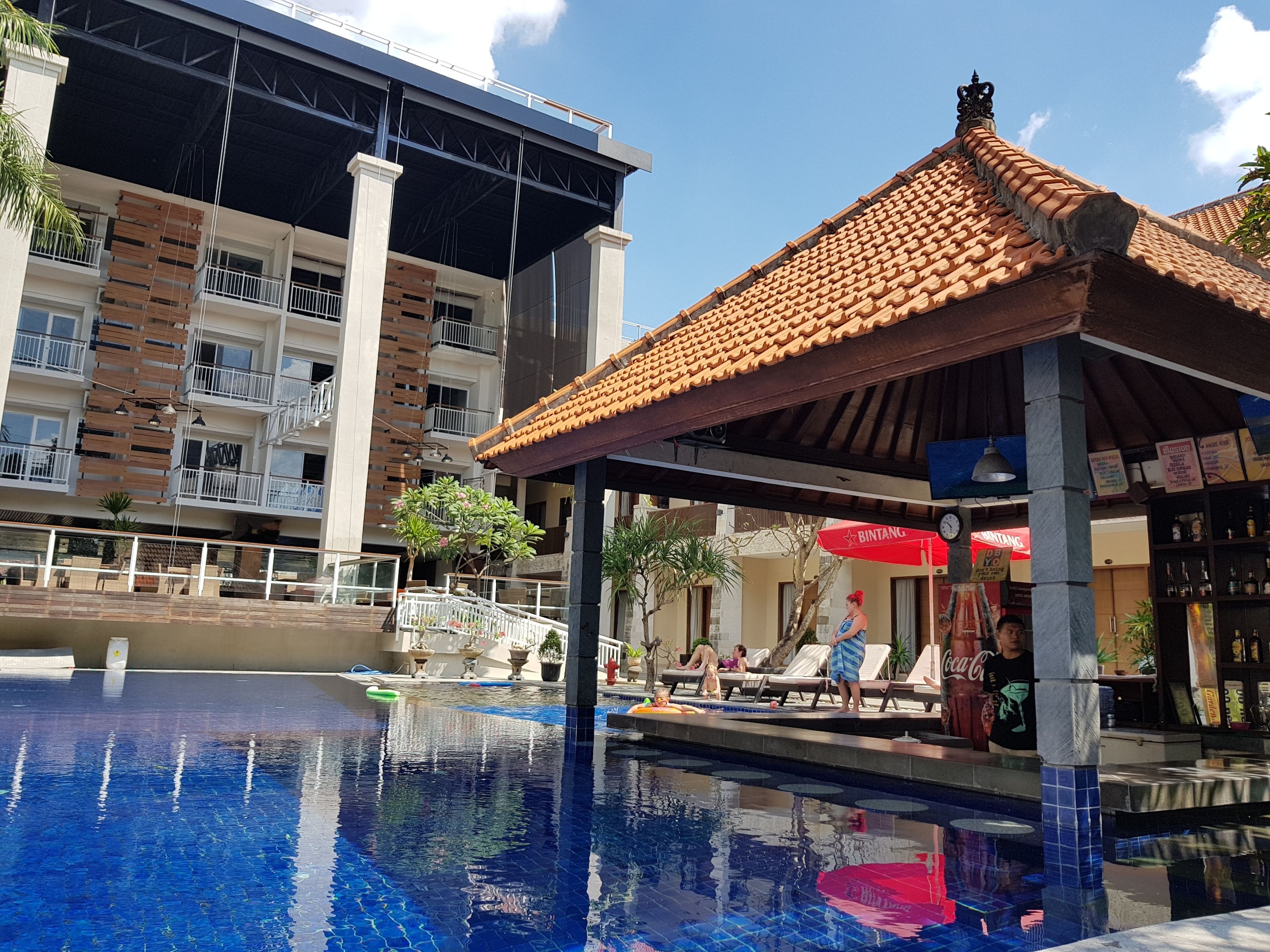 Una piscina con sillones de descanso y un área de estar cubierta, rodeada por un edificio moderno con balcones.