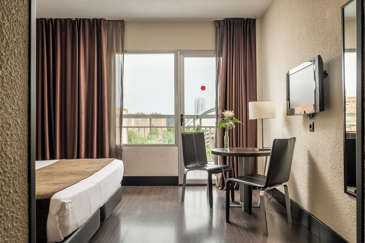 A hotel room with a bed, two chairs, a small table with a lamp, and a TV mounted on the wall.