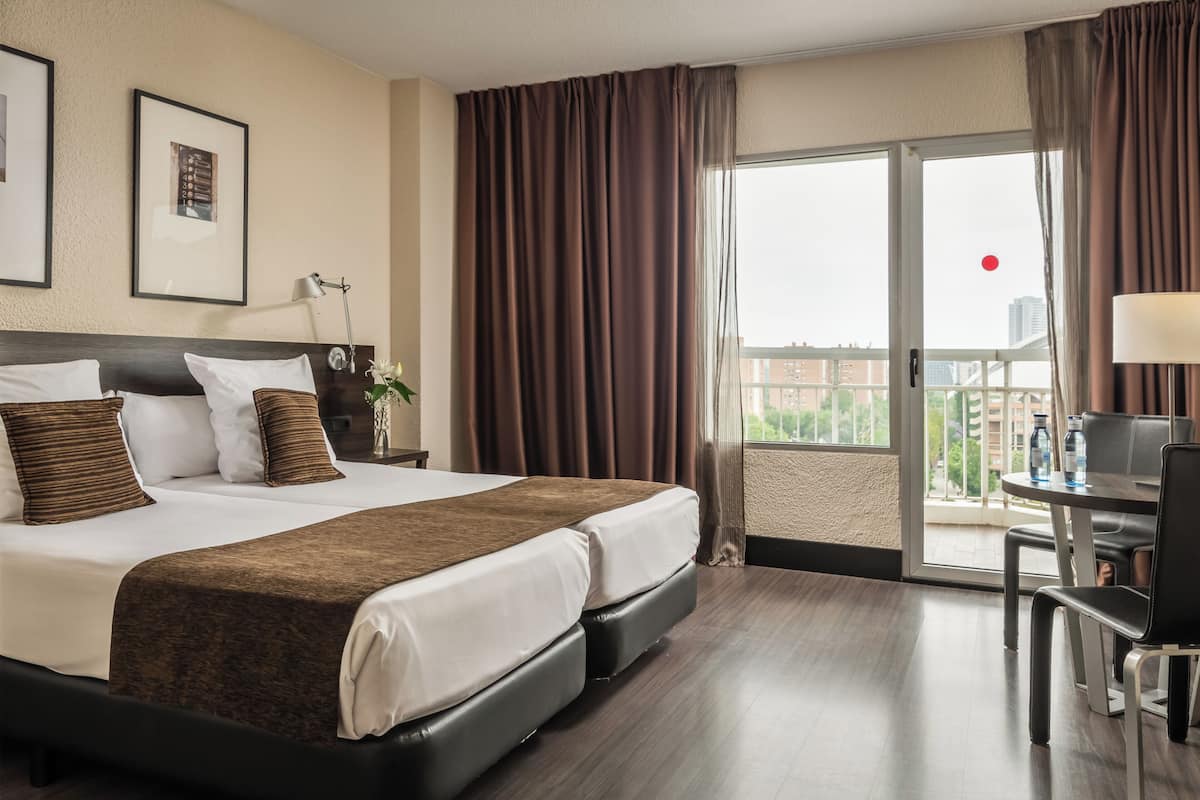 Ein Hotelzimmer mit einem großen Bett, einem Schreibtisch mit Stuhl, einem Balkon mit Vorhängen und Blick auf die Stadt.