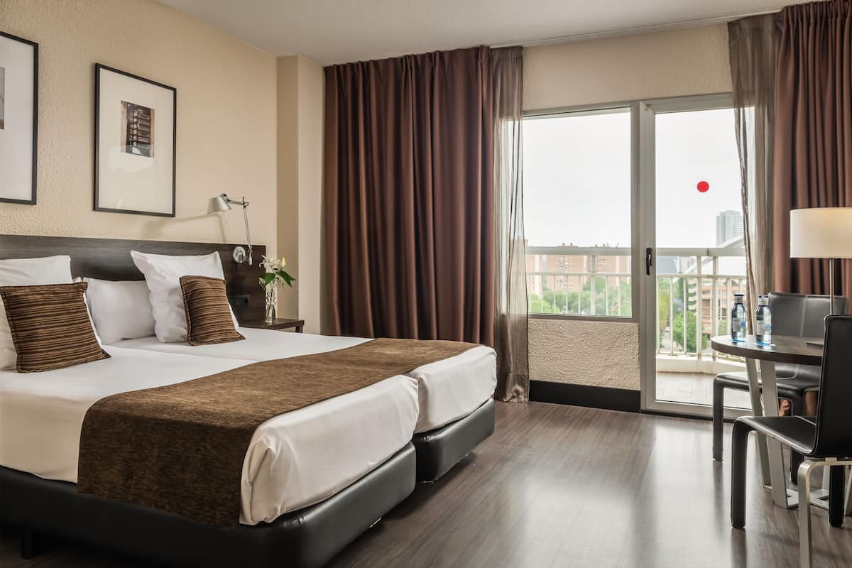 Ein Hotelzimmer mit einem großen Bett, einem Schreibtisch mit Stuhl, einem Balkon mit Vorhängen und Blick auf die Stadt.