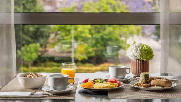 A breakfast table with a bowl of cereal, a glass of orange juice, a plate of fruit, a cup of coffee, a cup of tea, a small plant, and a pastry. - Hotel Medium Valencia (Valencia)
