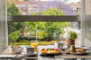 Een ontbijttafel met een kom ontbijtgranen, een glas sinaasappelsap, een bord fruit, een kop koffie, een kop thee, een kleine plant en een gebakje.