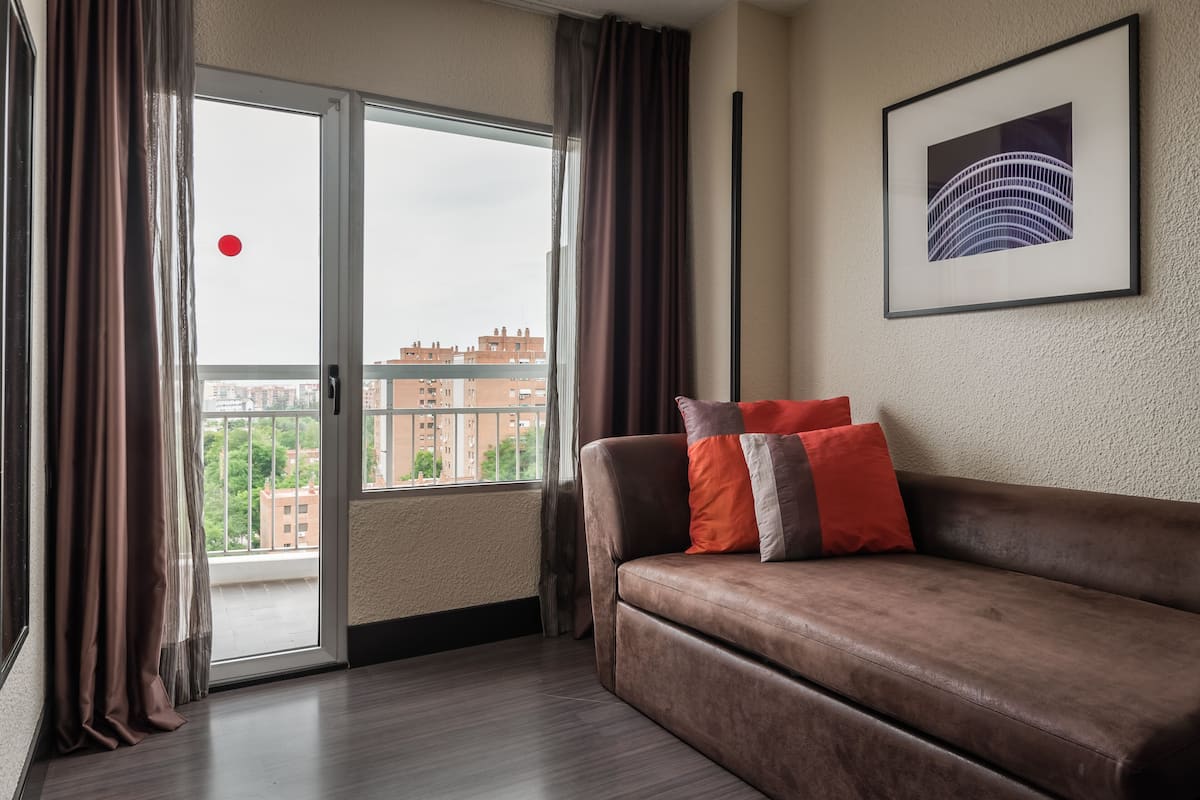 Ein modernes Wohnzimmer mit einem braunen Ledersofa, einem großen Fenster mit Vorhängen und einem gerahmten Bild an der Wand.