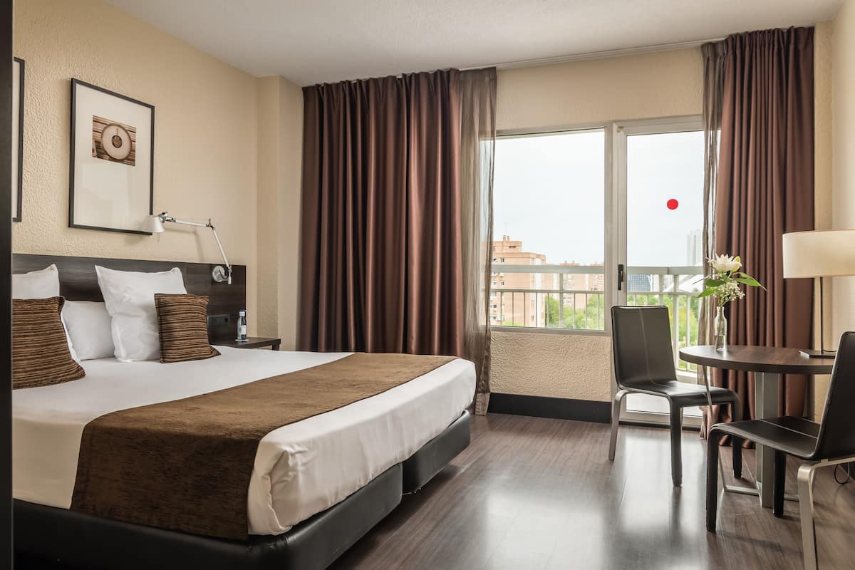 Ein Hotelzimmer mit einem großen Bett, einem Schreibtisch mit zwei Stühlen, einem Fenster mit Vorhängen und einem gerahmten Bild an der Wand.