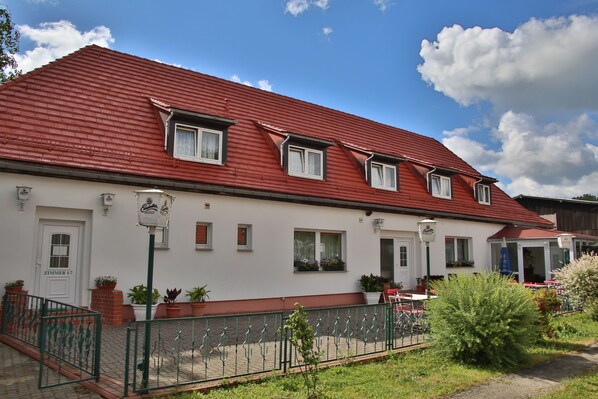 A two-story building with a red roof, white walls, and multiple windows. There is a small garden area with potted plants and a metal fence.