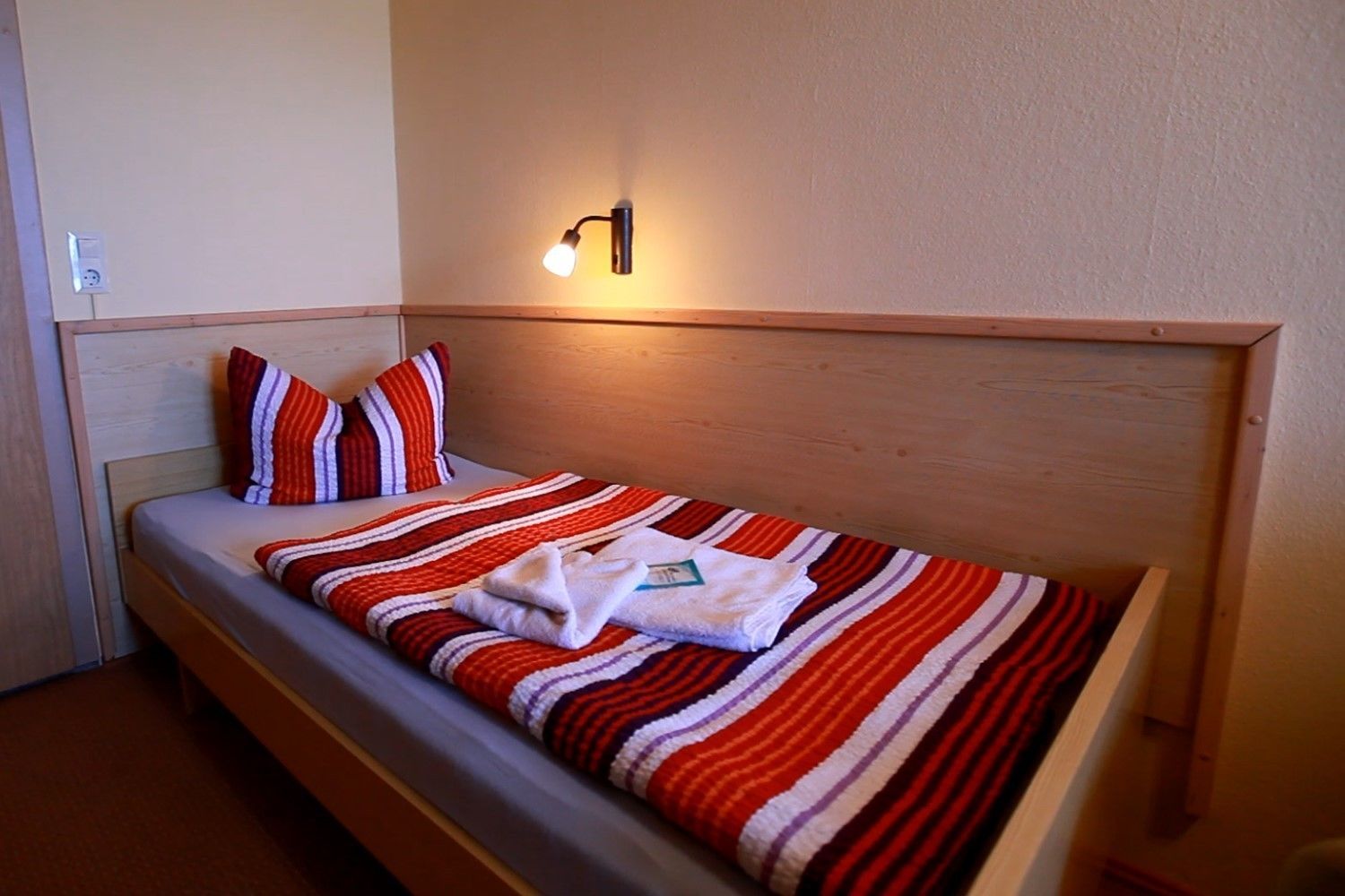 A single bed with a striped blanket, a pillow, and a folded towel. There is a wall-mounted light fixture above the bed.