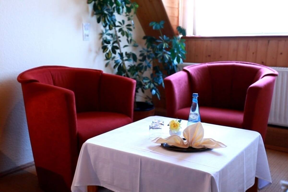 A room with two red armchairs, a small table with a white tablecloth, a bottle of water, and a decorative flower arrangement.