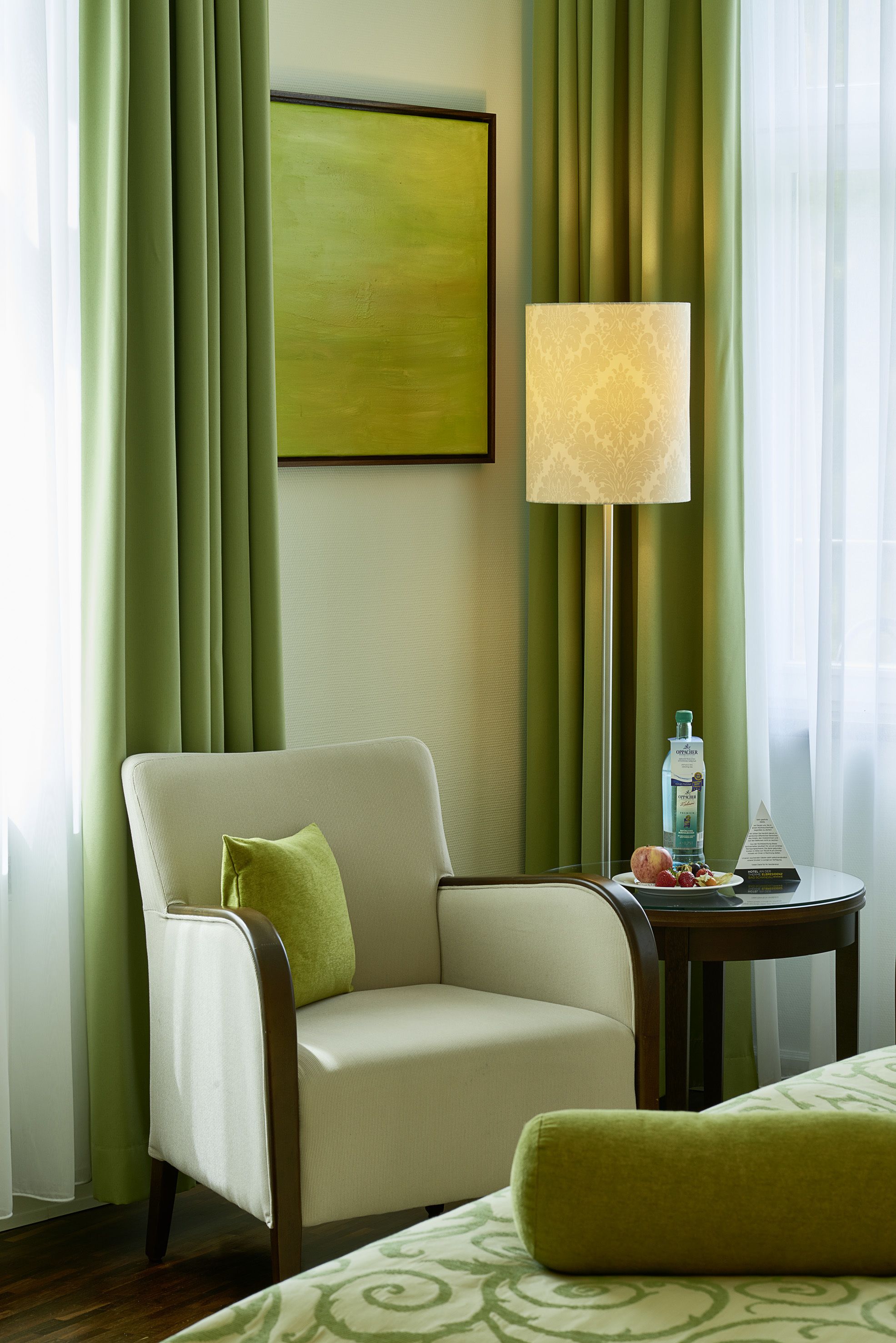 Habitación de hotel con decoración verde, que incluye cortinas, una silla con un cojín, una mesita de noche con una botella y fruta, y una cama con un almohada verde.