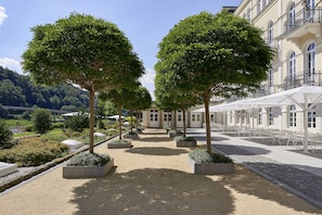 A paved walkway lined with trees and planters, leading to a building with outdoor seating.