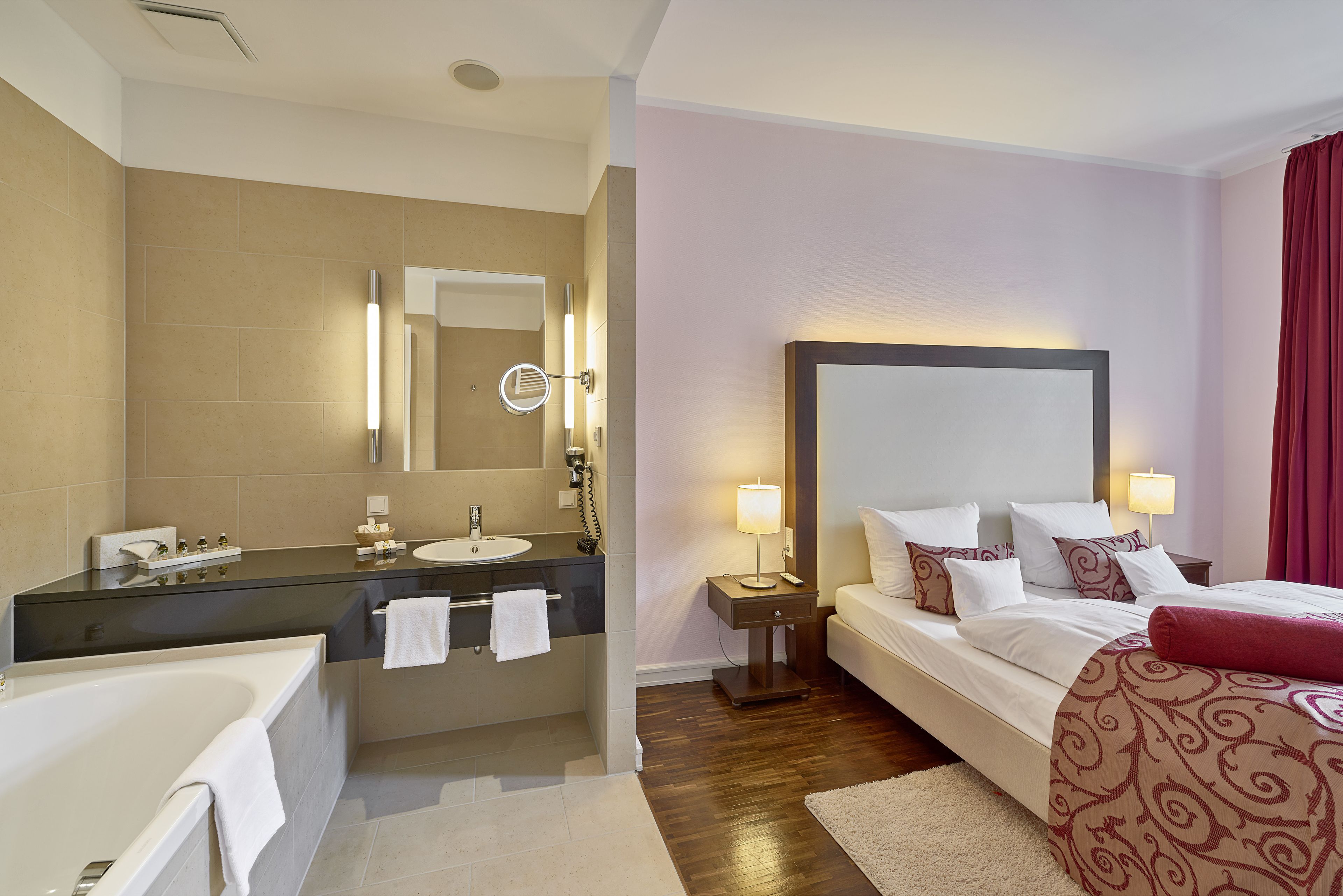 Una habitación de hotel con bañera, una cama grande con almohadas, una mesita de noche con lámpara y un espejo sobre el lavabo.