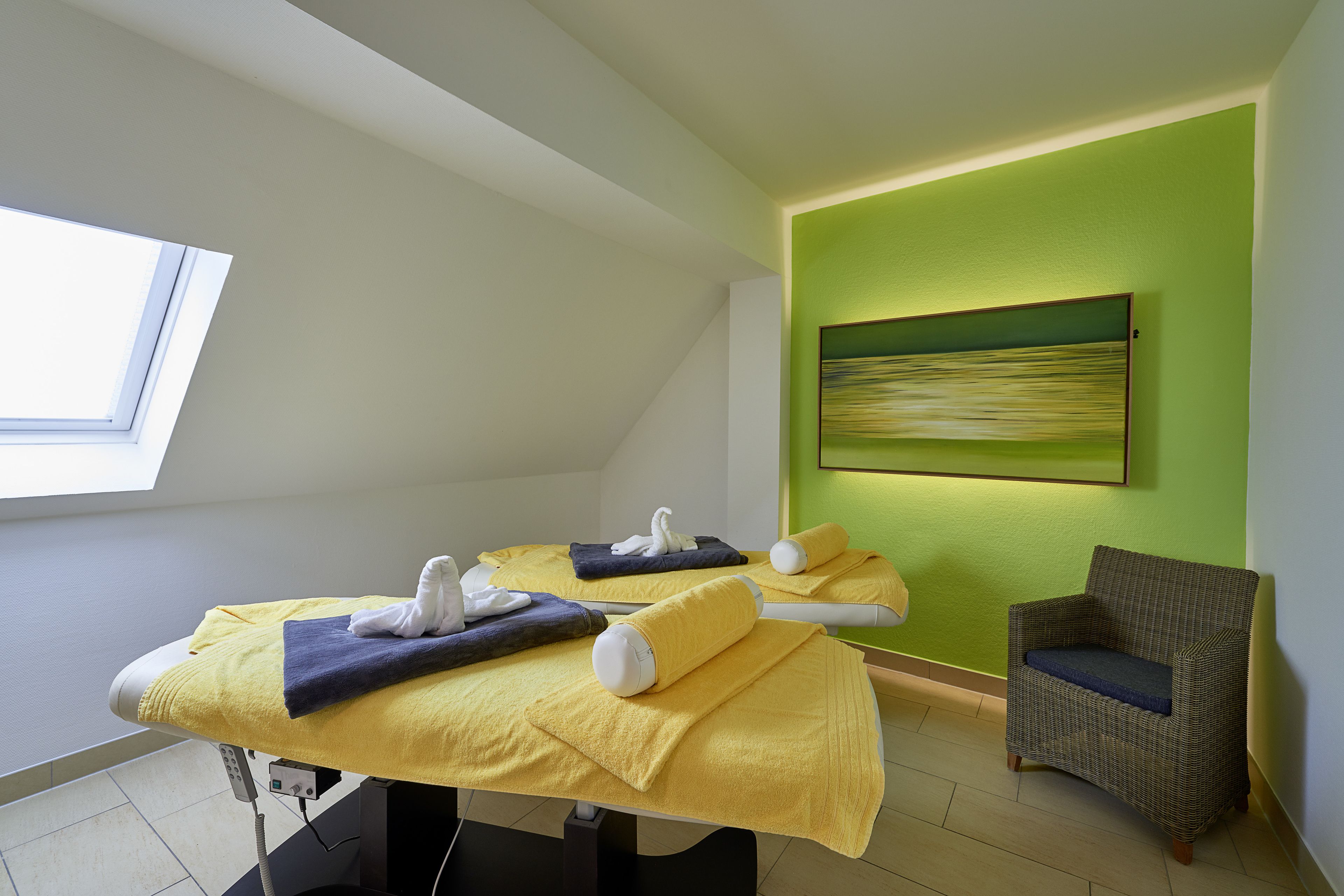 Un cuarto de tratamientos con dos camillas de masajes, una silla y un cuadro en la pared.