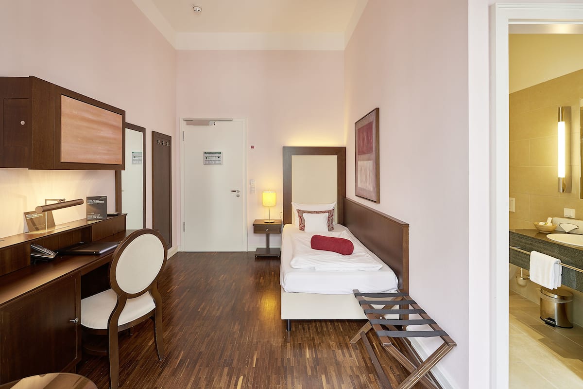 A hotel room with a wooden desk, a chair, a bed with white bedding, a bedside table with a lamp, and a bathroom with a sink and mirror.