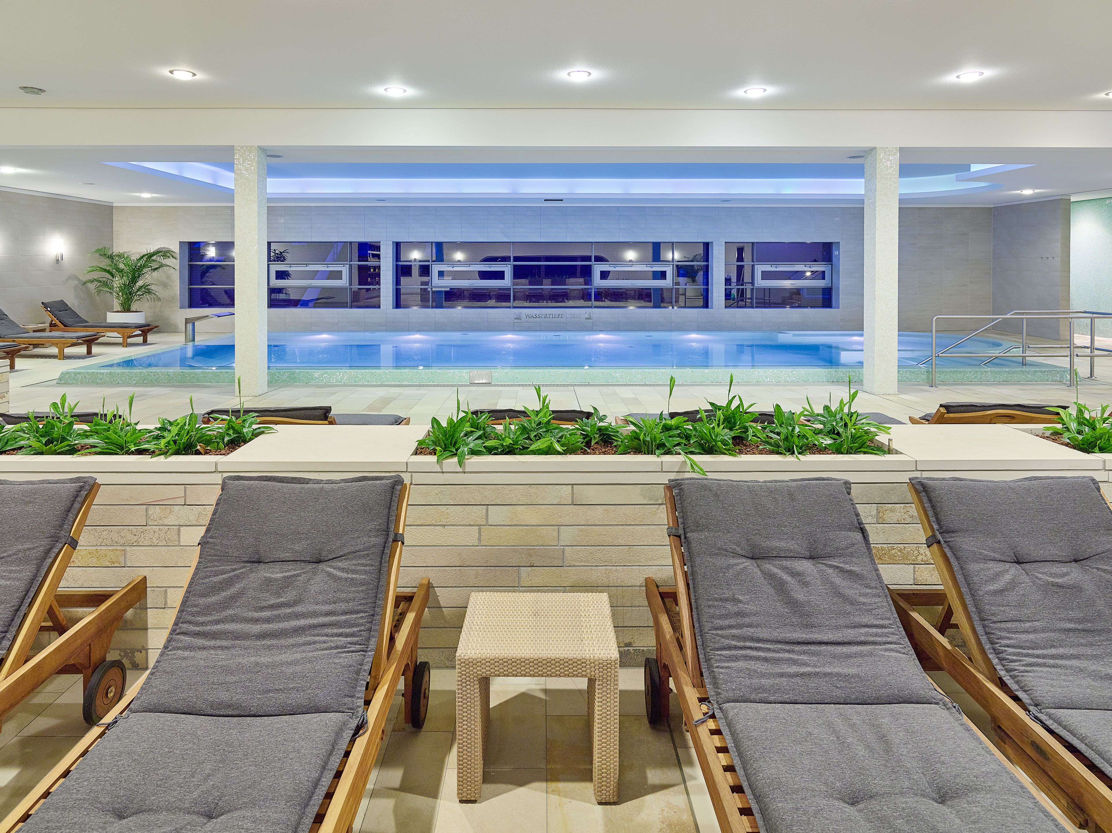 Zona de piscina con sillas de descanso y una pequeña mesa, rodeada de plantas.