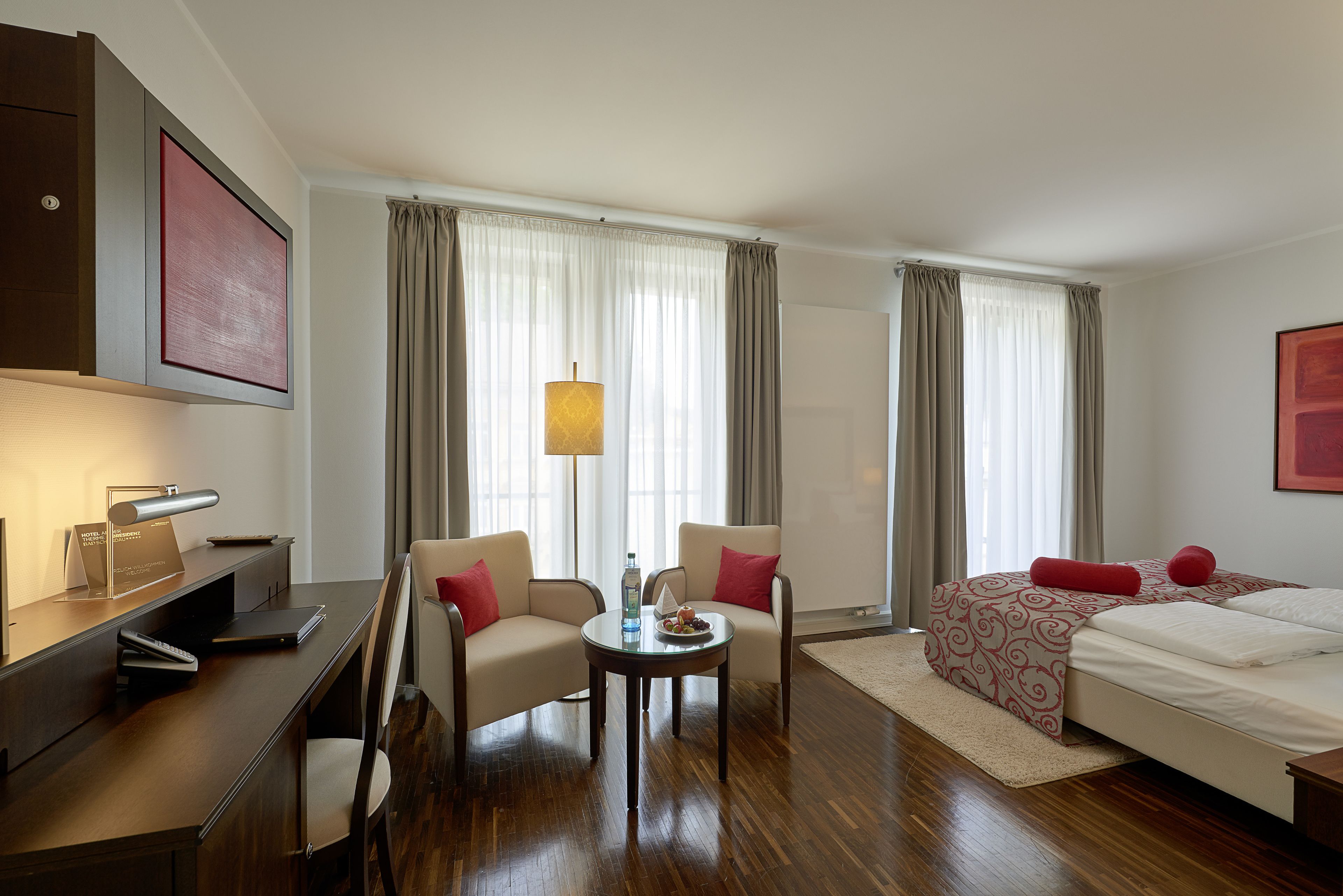 Una habitación de hotel con escritorio, dos sillones, una cama con colcha estampada, una lámpara de pie y un ventanal grande con cortinas.