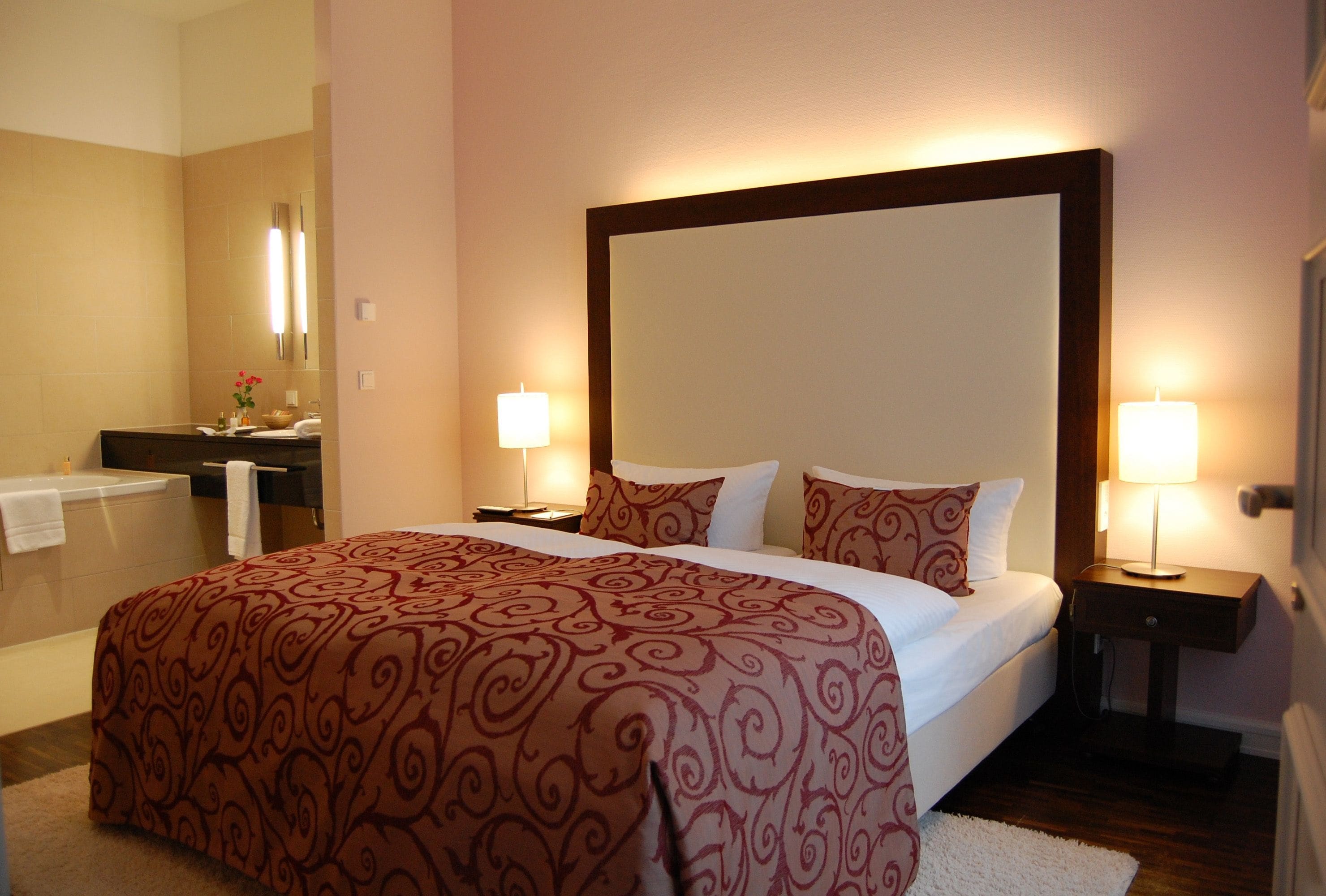 A hotel room with a large bed, two bedside tables with lamps, a nightstand, a bathroom with a bathtub, and a mirror.