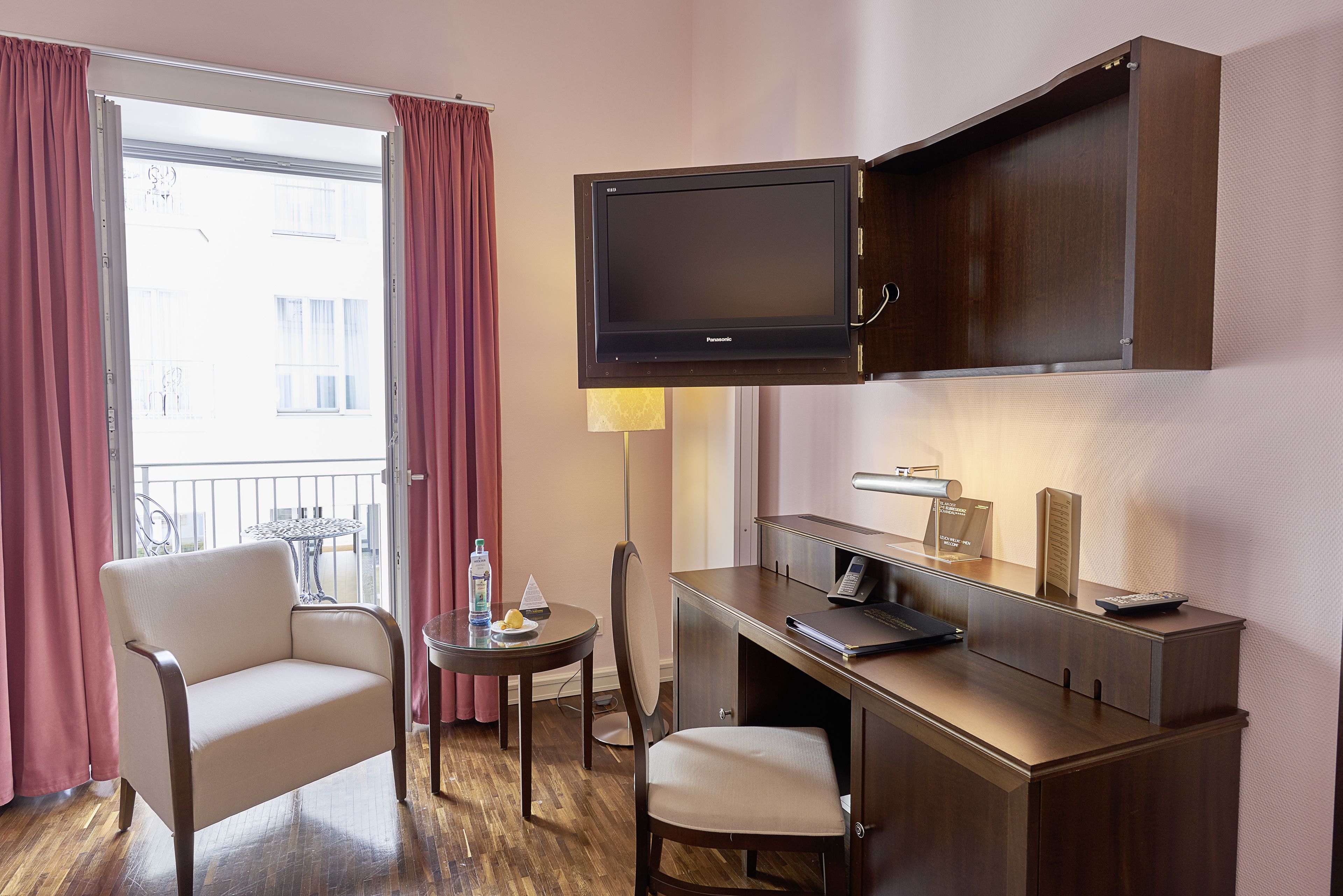 Habitación de hotel con escritorio, silla, televisor y vistas al balcón exterior.