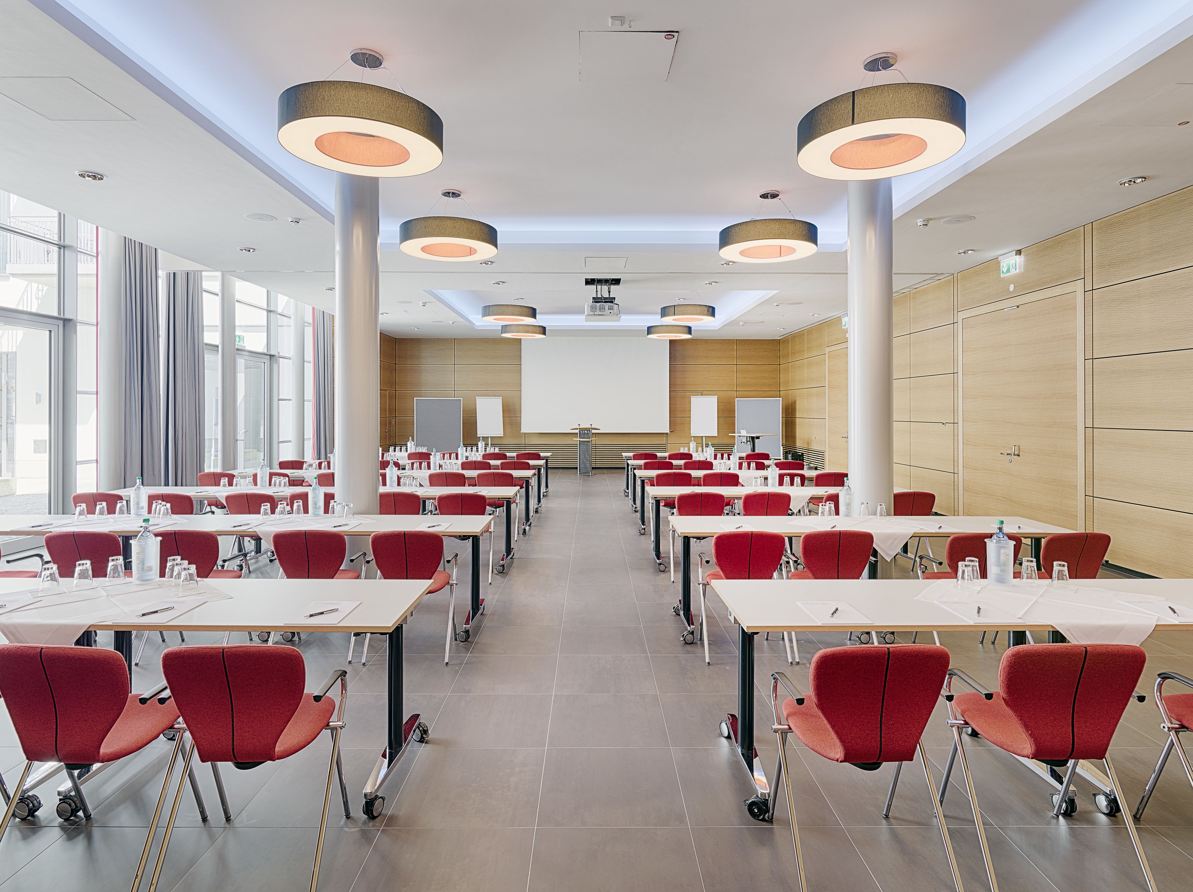 Un salón de conferencias con filas de mesas y sillas, ventanas amplias y luminarias modernas.