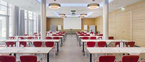 Un salón de conferencias con filas de mesas y sillas, ventanas amplias y luminarias modernas.