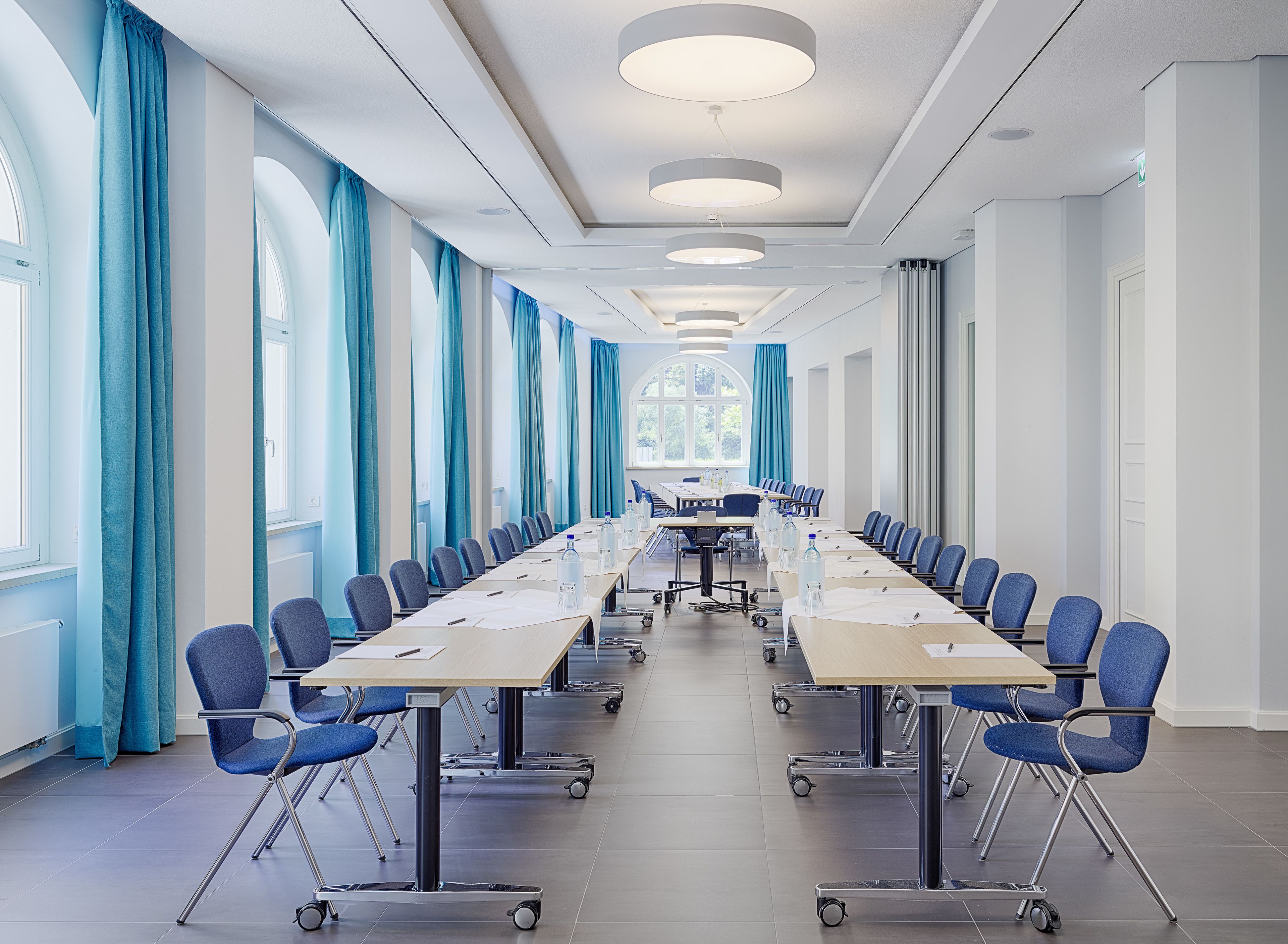 A conference room with a long table, blue chairs, and a view of the outdoors through large windows.