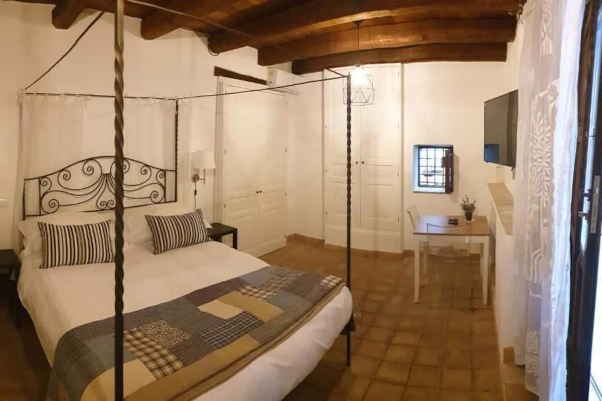 Ein Bett mit Baldachin, eine Bettdecke, Nachttische, ein Schreibtisch mit Stuhl und ein Fernseher an der Wand.