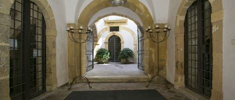 Un corredor arqueado con paredes de piedra y puertas de metal, que conduce a un patio con plantas en macetas.