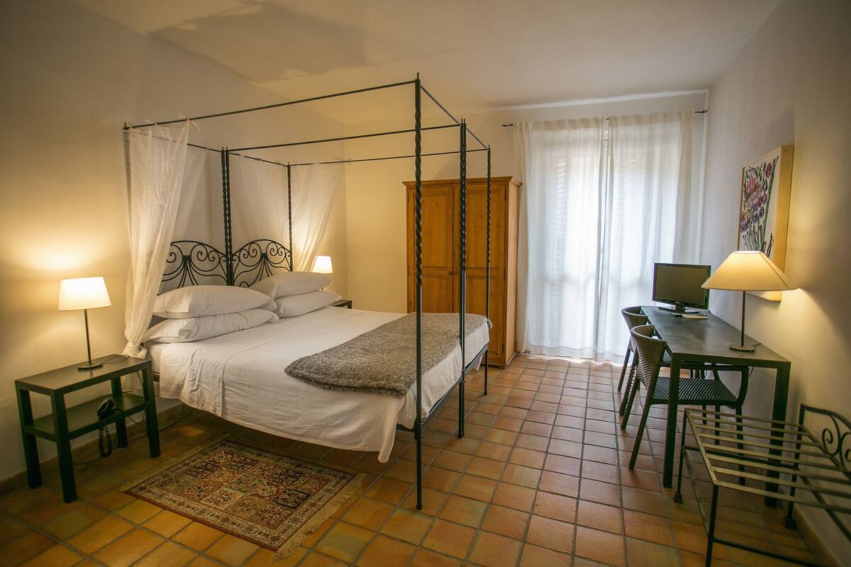 Ein Bett mit Baldachin, ein Holzkopfteil, ein Schreibtisch mit Stuhl und ein kleiner Tisch mit Lampe.