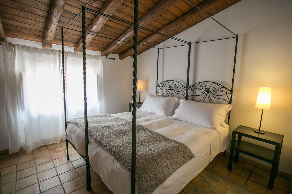 Ein Bett mit Himmelbett, ein Holz-Kopfteil, ein Nachttisch mit Lampe und ein Fenster mit Vorhängen.
