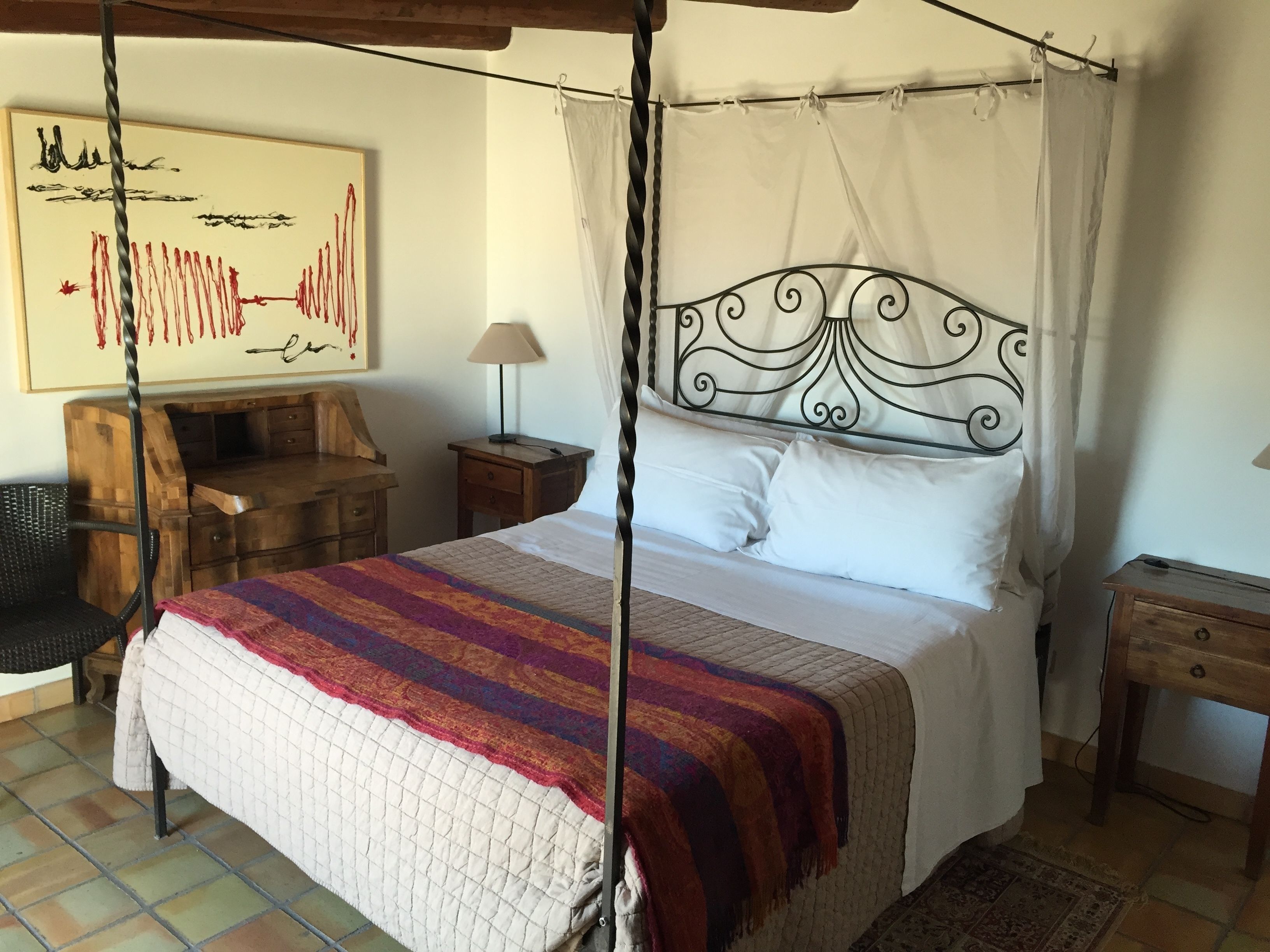 A four-poster canopy bed with white linens, a colorful striped blanket, and a headboard with intricate metalwork. There is a wooden nightstand with a lamp, a wooden desk, and a chair to the left. A framed abstract painting is on the wall above the bed.