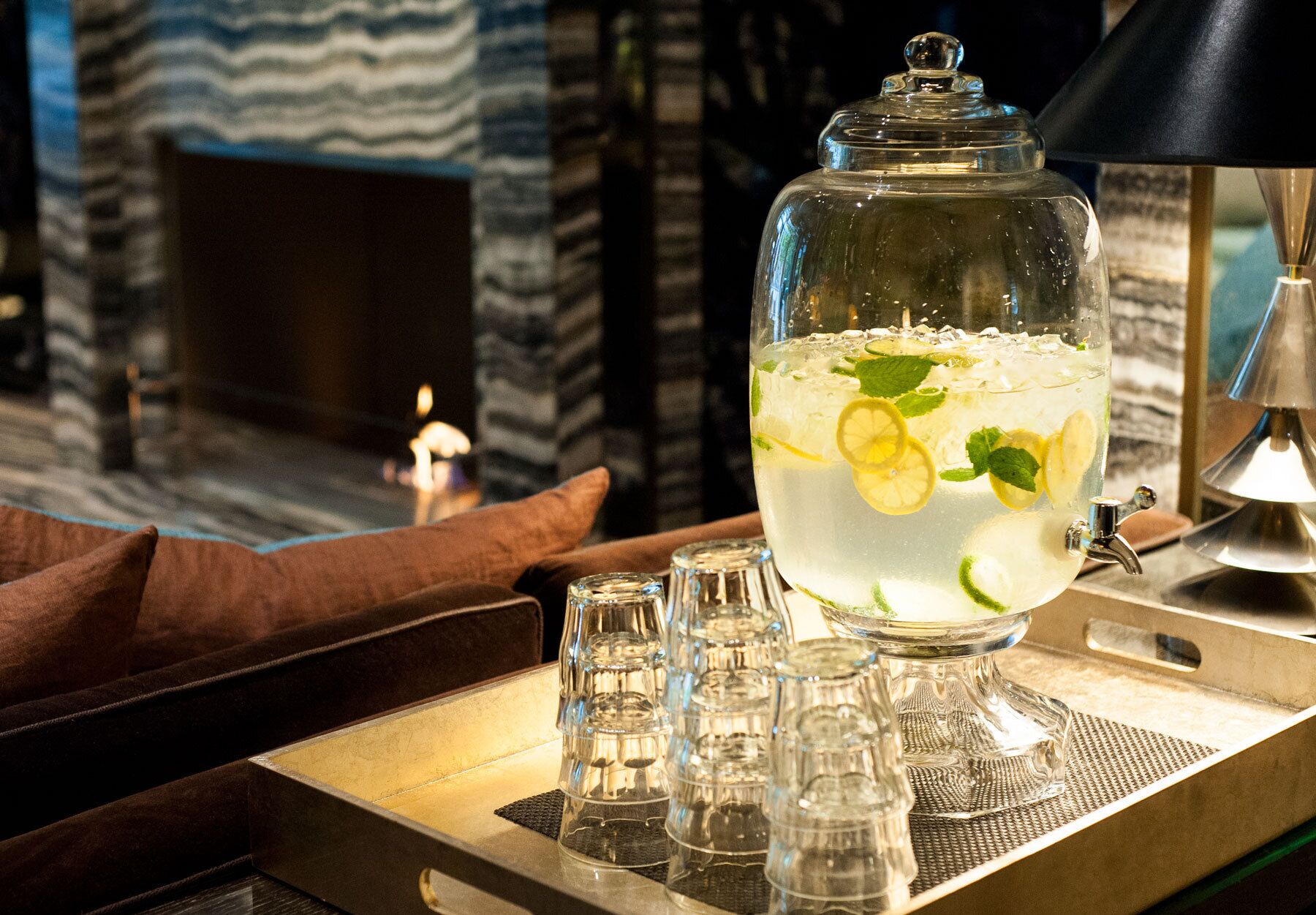 A clear glass pitcher filled with a beverage, garnished with lemon slices and mint leaves, placed on a tray with a candle and salt and pepper shakers.