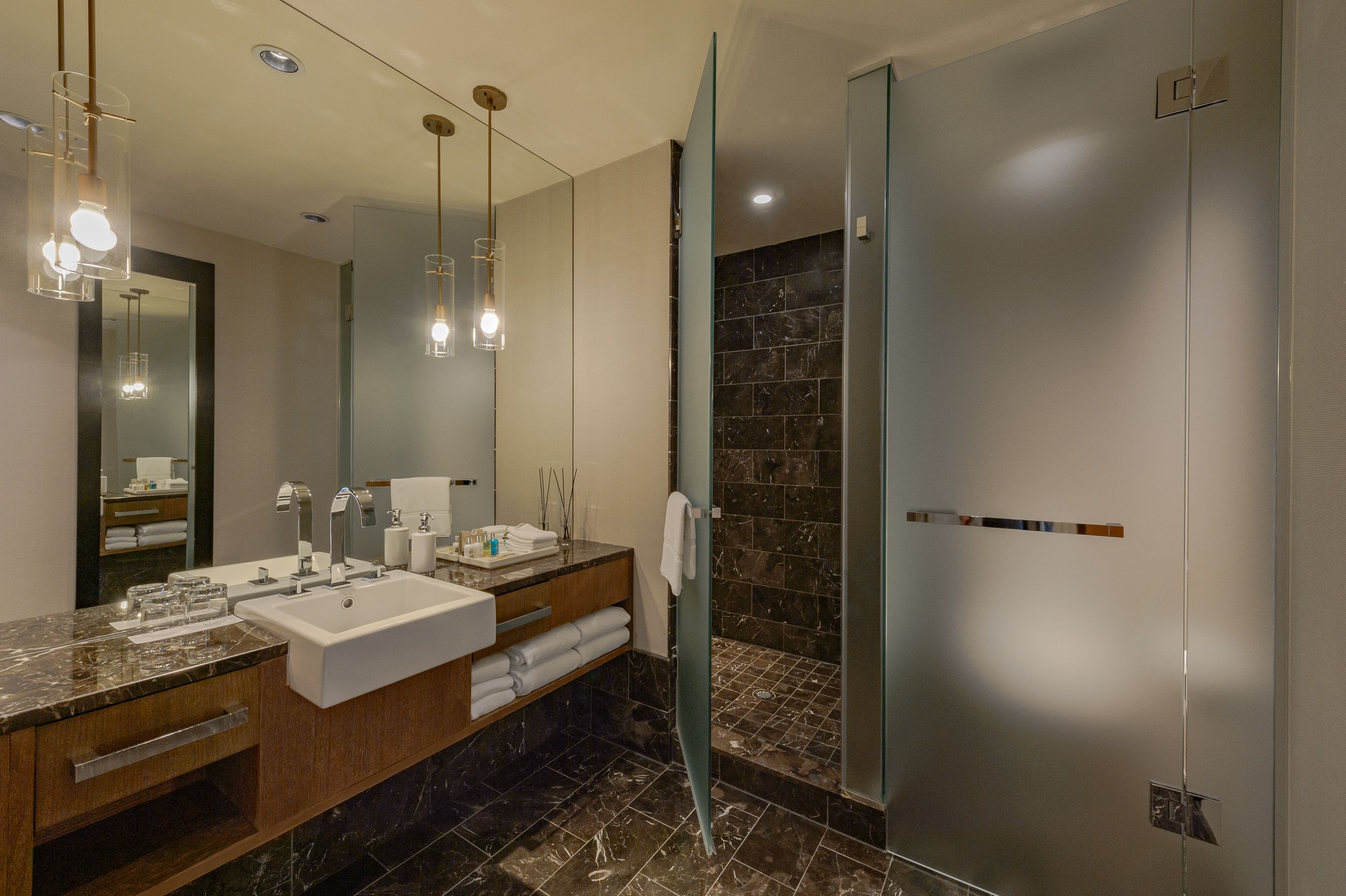 A modern bathroom with a large mirror, two sinks, a glass shower enclosure, and a marble-tiled floor.