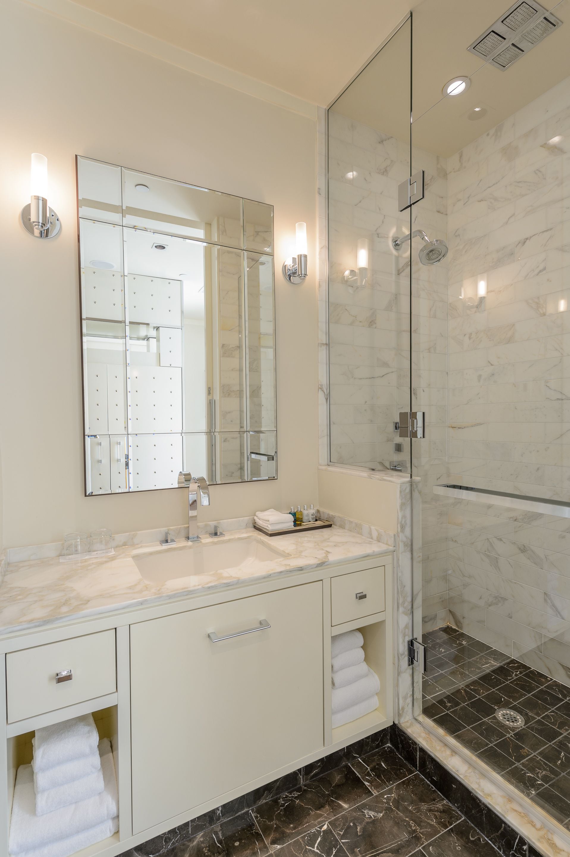 A modern bathroom with a large mirror, a glass-enclosed shower, and marble flooring.
