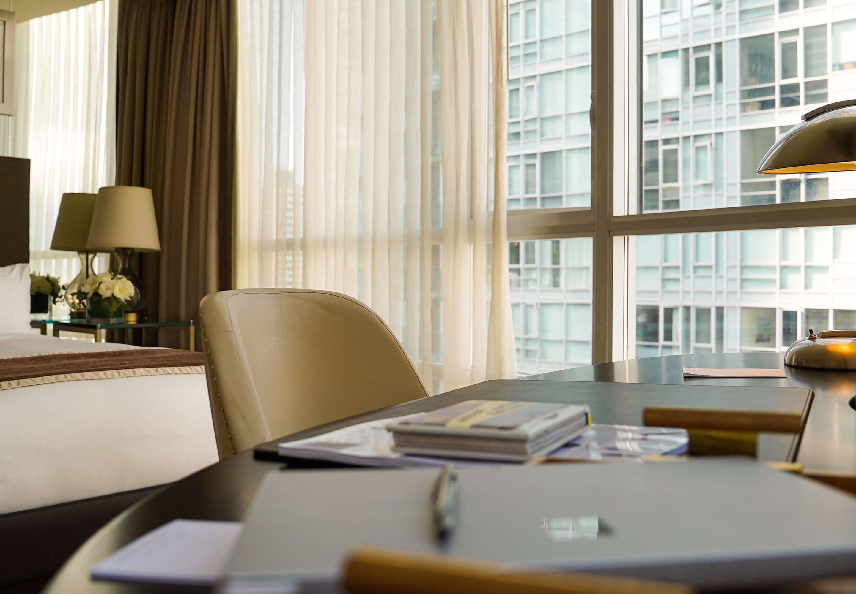 A hotel room with a bed, a desk with a lamp, a chair, and a view of a building through the window.